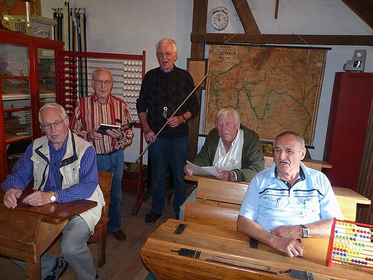 „Alle Vöglein sind schon da!“ Beim Rundgang durch die Ausstellung nehmen die fleißigen Helfer auf den alten Schulbänken Platz, Günther Birkle schwingt beim gemeinsamen Lied den (Takt-)-Stock.