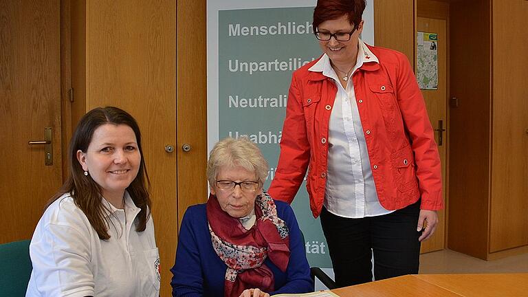 Katja Simon, Fahrerin beim &bdquo;Essen auf Rädern des BRK Rhön-Grabfeld&rdquo; (links) und Nicole Herbart vom BRK Sozialdienst, blättern mit einem Tischgast im Menü-Katalog