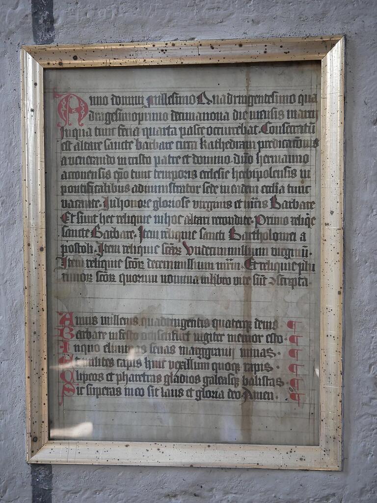 Eingelassen in eine Säule und geschützt durch eine Glasscheibe und einem Metallrahmen hat die Weiheinschrift des Barbara-Altars aus dem 1441 bis heute überdauert.
