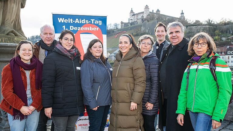 Veronika Schmid (v.l.), Dieter Wenderlein, Julia Hemmrich, Heidrun Brand, Hülya Düber, Monika Kraft, David Hein, Domkapitular Clemens Bieber und Brigitte Hein stehen auf der Alten Mainbrücke in Würzburg vor der Aids-Schleife am Festungsberg.