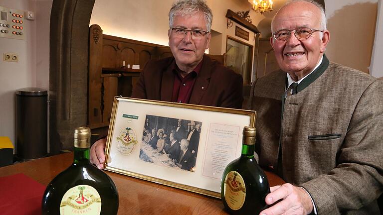 Der langjährige Bürgerspital-Weingutsleiter Rudolf Frieß (rechts) mit seinem Nachfolger Robert Haller&nbsp; im Jahr 2015. Im Vordergrund zwei Flaschen des Weins für das Essen mit der Queen 1965. Das Foto im goldenen Rahmen zeigt die Queen beim Essen auf Schloss Charlottenburg am 27. Mai 1965. Mit eingerahmt sind auch das Etikett des Bürgerspital-Weins und die Speisekarte.