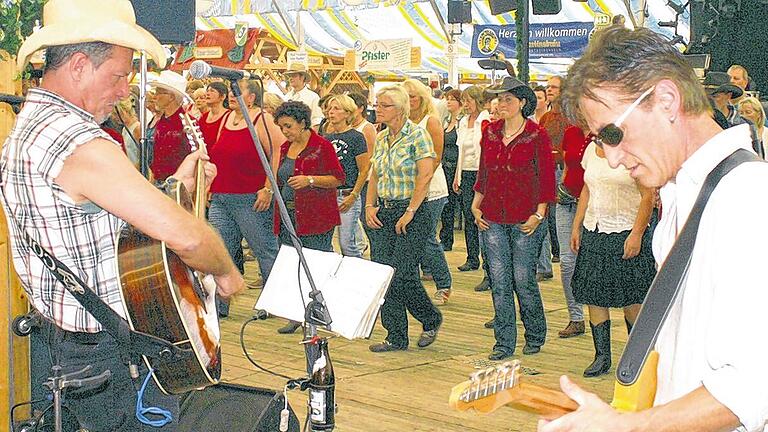 Ein Tänzchen zum Westernfrühschoppen       -  (maha)   Jeans, Karohemden, Boots und Cowboyhüte prägten am Sonntagmorgen wieder das Bild des Laurenzi-Festzelts in Marktheidenfeld, denn seit einigen Jahren ist der Westernfrühschoppen zu einem beliebten Treffpunkt der Fans US-amerikanischer Country-Musik am Main geworden. Dazu werden einige Tischreihen vor der Bühne auf Seite geräumt, um einer recht beachtlichen Schar von Line-Dancern Platz für ihre für jedermann choreografierten Tanzeinlagen zu bieten. In diesem Jahr waren wieder die fünf Musiker der beliebten Band &bdquo;Country Blend&ldquo; zu Gast. Mit Fiddle, Steelguitar, Gitarre, Bass, Schlagzeug und weiteren Instrumenten spielte das Quintett versiert, was die Szene schätzt &ndash; vom Traditional, über den amerikanischen Roots-Rock der Südstaaten bis hin zu den unvergesslichen John-Fogerty-Hits und der Band Creedence Clearwater Revival. In die Tanzformationen reihten sich auch gerne einige unerfahrene Gäste ein und wagten ein paar erste Schritte. So wurde der Morgen wieder zu einem Vergnügen für alt und jung, groß und klein, schlank oder korpulent &ndash; einfach für jeden!