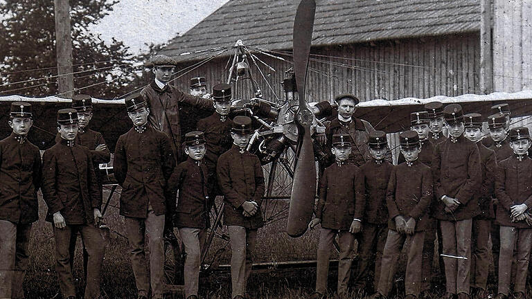 Lendner-Eindecker im September 1912: Der Würzburger Flugpionier Leo Lendner (mit Zivilmütze) steht erhöht auf einem der Fahrwerksräder. Davor sind k.u.k.-österreichische Militärkadetten, zu Besuch auf dem Exerzierplatz Kugelfang am Galgenberg, zu sehen, Würzburgs damaligem provisorischem Flugplatz. Einer von ihnen durfte einen Flug mit Lendner machen und hat dazu bereits im &bdquo;Aeroplan&ldquo; Platz genommen. Im Hintergrund Würzburgs erste Flugzeughalle, in der Lendner das Flugzeug selbst gebaut hatte.