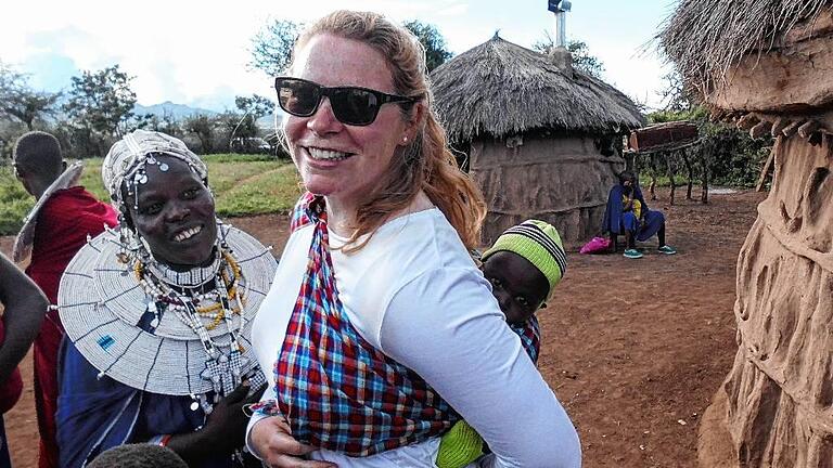 Susanne Wolf trägt das Baby einer Maasai-Mutter, die daneben steht, in einem typischen Maasai-Dorf in Tansania.