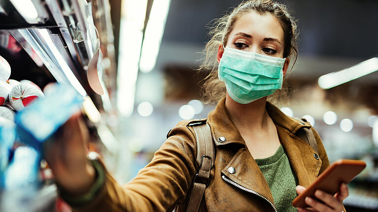 Woman wearing protective mask while using cell phone and buying food in grocery store during virus epidemic.       -  Regeln und Empfehlungen zur Eindämmung des Coronavirus sind im öffentlichen Raum nicht immer einfach einzuhalten.