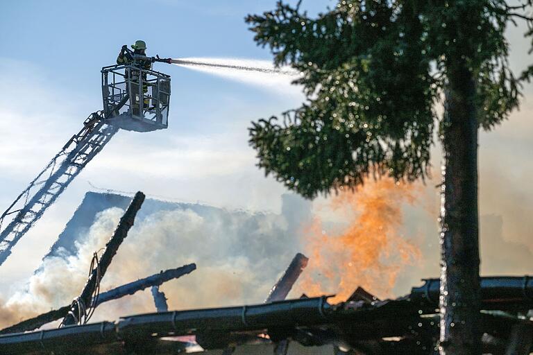 Ein Feuerwehrmann strahlt bei dem Brand in Pfaffendorf mit Wasser von einer Drehleiter, während das Gebälk einer Scheune bereits komplett verkohlt ist und Flammen aus ihr herausschlagen. Ein Feuerwehrmann strahlt bei dem Brand in Pfaffendorf mit Wasser von einer Drehleiter, während das Gebälk einer Scheune bereits komplett verkohlt ist und Flammen aus ihr herausschlagen.