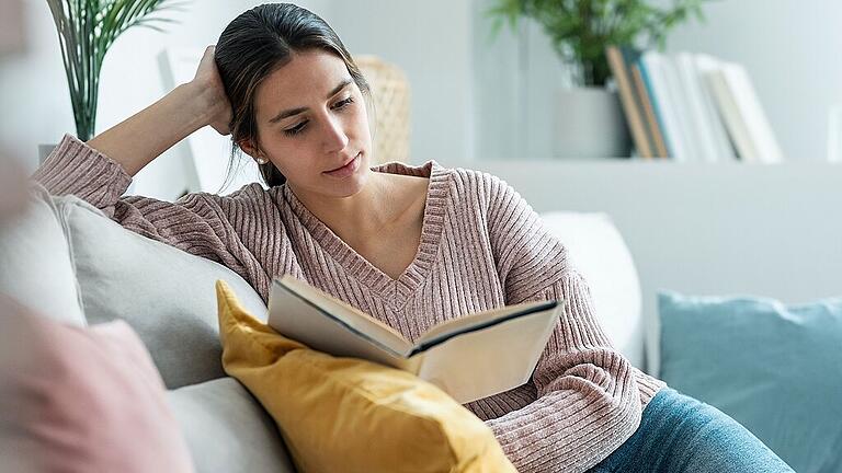 Pretty young woman reading a book while sitting on sofa at home.       -  Mit einem guten Buch in der Hand kann man im Herbst bestens auf der Couch entspannen.