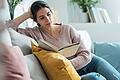 Pretty young woman reading a book while sitting on sofa at home.       -  Mit einem guten Buch in der Hand kann man im Herbst bestens auf der Couch entspannen.